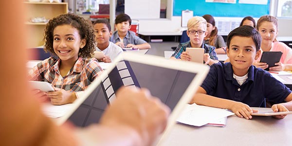 Students sitting in a classroom looking at their teacher holding a tablet